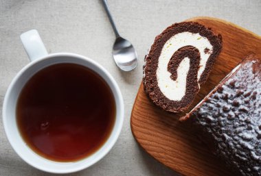 Çikolatalı sünger rulo vanilya kremalı tahta tahtada bir fincan çayın yanında.