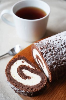 Çikolatalı sünger rulo vanilya kremalı tahta tahtada bir fincan çayın yanında.