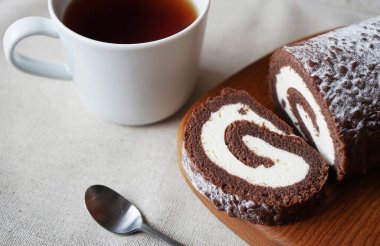 Çikolatalı sünger rulo vanilya kremalı tahta tahtada bir fincan çayın yanında.