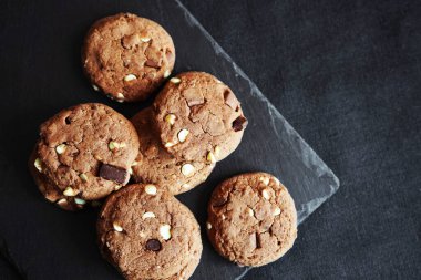 Siyah bir arka planda fındıklı siyah çikolata dilimlerinin yanında çikolatalı kurabiyeler.