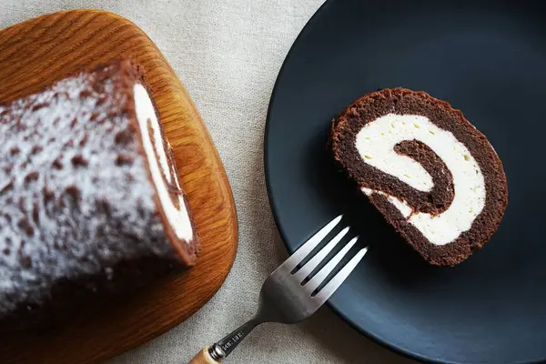Tahtada vanilya kremalı çikolatalı dürüm, arka planda açık bir tabakta kesilmiş ruloların yanında.