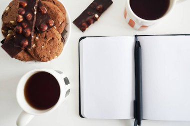 Çikolatalı kurabiyeli iki fincan çay ve beyaz arka planda açık bir not defterinin yanında fındıklı çikolata.