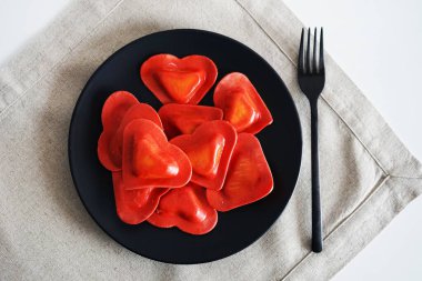 Siyah tabakta kırmızı kalp şeklinde ravioli, arka planda bir çatalın yanında.