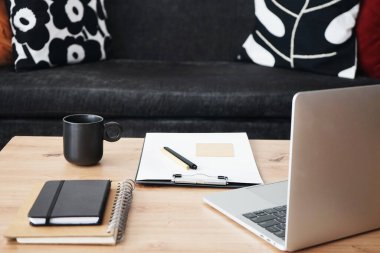 Açık dizüstü bilgisayarı, klasörleri, kalemi ve siyah kupası olan gri yastıklı kanepenin arka planında kahve olan işyeri.