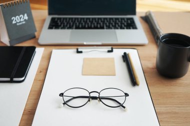 Açık dizüstü bilgisayarı, dizinleri, klasörleri, kalemi, gözlükleri, takvimi ve kahveli siyah kupası olan işyeri