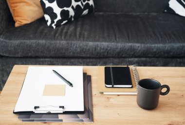 Not defterleri, klasörleri, kalemi ve siyah kupası olan gri yastıklı kanepenin arka planında kahve olan bir masaüstü.