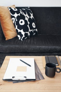 Not defterleri, klasörleri, kalemi ve siyah kupası olan gri yastıklı kanepenin arka planında kahve olan bir masaüstü.