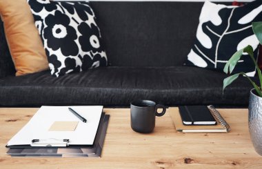 Not defterleri, klasörleri, kalemi ve siyah kupası olan gri yastıklı kanepenin arka planında kahve olan bir masaüstü.