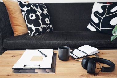 Not defterleri, klasörleri, kalemleri, kulaklıkları, takvimi ve arka planda yastıklı gri kanepenin kahvesi olan siyah kupası olan masaüstü.