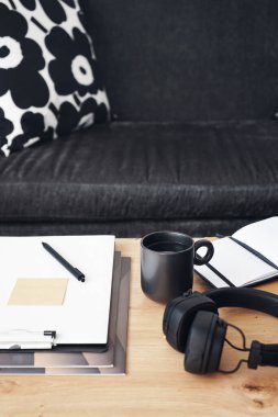 Not defterleri, klasörleri, kalemleri, kulaklıkları, takvimi ve arka planda yastıklı gri kanepenin kahvesi olan siyah kupası olan masaüstü.