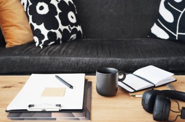 Not defterleri, klasörleri, kalemleri, kulaklıkları, takvimi ve arka planda yastıklı gri kanepenin kahvesi olan siyah kupası olan masaüstü.