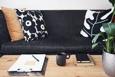 Not defterleri, klasörleri, kalemi ve siyah kupası olan gri yastıklı kanepenin arka planında kahve olan bir masaüstü.