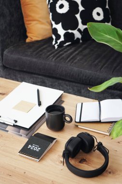 Not defterleri, klasörleri, kalemleri, kulaklıkları, takvimi ve arka planda yastıklı gri kanepenin kahvesi olan siyah kupası olan masaüstü.