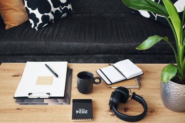 Not defterleri, klasörleri, kalemleri, kulaklıkları, takvimi ve arka planda yastıklı gri kanepenin kahvesi olan siyah kupası olan masaüstü.