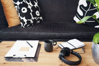 Not defterleri, klasörleri, kalemleri, kulaklıkları, takvimi ve arka planda yastıklı gri kanepenin kahvesi olan siyah kupası olan masaüstü.