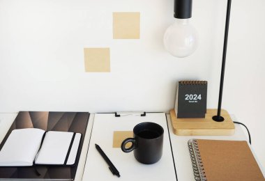 Not defterleri, klasörler, çıkartmalar, takvim, bardaklar, kalemler, masa lambası açık arkaplanda siyah bir fincan kahvenin yanında.