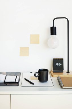 Not defterleri, klasörler, çıkartmalar, takvim, bardaklar, kalemler, masa lambası açık arkaplanda siyah bir fincan kahvenin yanında.