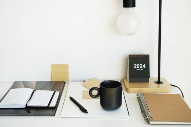 Not defterleri, klasörler, çıkartmalar, takvim, bardaklar, kalemler, masa lambası açık arkaplanda siyah bir fincan kahvenin yanında.