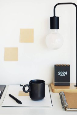 Not defterleri, klasörler, çıkartmalar, takvim, bardaklar, kalemler, masa lambası açık arkaplanda siyah bir fincan kahvenin yanında.