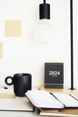 Not defterleri, klasörler, çıkartmalar, takvim, bardaklar, kalemler, masa lambası açık arkaplanda siyah bir fincan kahvenin yanında.