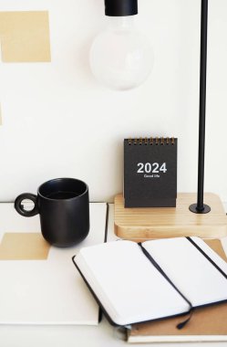 Not defterleri, klasörler, çıkartmalar, takvim, bardaklar, kalemler, masa lambası açık arkaplanda siyah bir fincan kahvenin yanında.