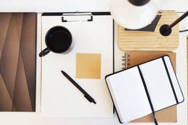 Not defterleri, klasörler, çıkartmalar, takvim, bardaklar, kalemler, masa lambası açık arkaplanda siyah bir fincan kahvenin yanında.