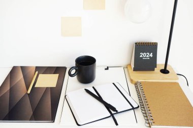 Not defterleri, klasörler, çıkartmalar, takvim, bardaklar, kalemler, masa lambası açık arkaplanda siyah bir fincan kahvenin yanında.