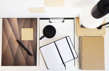 Not defterleri, klasörler, çıkartmalar, takvim, bardaklar, kalemler, masa lambası açık arkaplanda siyah bir fincan kahvenin yanında.