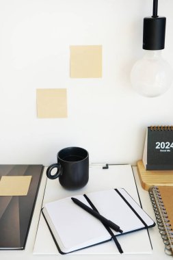 Not defterleri, klasörler, çıkartmalar, takvim, bardaklar, kalemler, masa lambası açık arkaplanda siyah bir fincan kahvenin yanında.