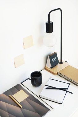 Not defterleri, klasörler, çıkartmalar, takvim, bardaklar, kalemler, masa lambası açık arkaplanda siyah bir fincan kahvenin yanında.