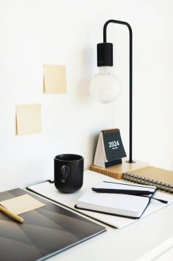 Not defterleri, klasörler, çıkartmalar, takvim, bardaklar, kalemler, masa lambası açık arkaplanda siyah bir fincan kahvenin yanında.