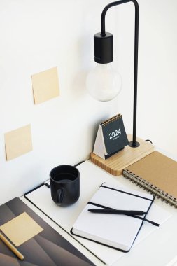 Not defterleri, klasörler, çıkartmalar, takvim, bardaklar, kalemler, masa lambası açık arkaplanda siyah bir fincan kahvenin yanında.