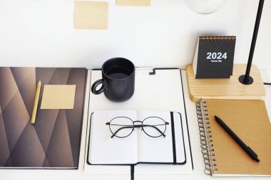 Not defterleri, klasörler, çıkartmalar, takvim, bardaklar, kalemler, masa lambası açık arkaplanda siyah bir fincan kahvenin yanında.