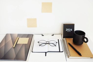 Not defterleri, klasörler, çıkartmalar, takvim, bardaklar, kalemler açık arkaplanda siyah bir fincan kahvenin yanında.