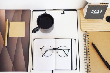 Not defterleri, klasörler, çıkartmalar, takvim, bardaklar, kalemler, masa lambası açık arkaplanda siyah bir fincan kahvenin yanında.