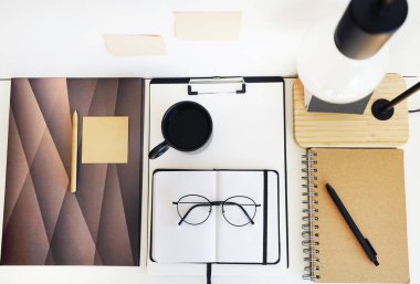 Not defterleri, klasörler, çıkartmalar, takvim, bardaklar, kalemler, masa lambası açık arkaplanda siyah bir fincan kahvenin yanında.