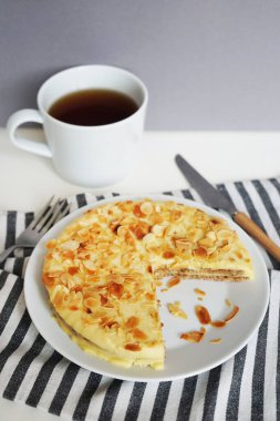 Beyaz bir tabakta bademli kek çizgili bir havlunun üzerinde, çatal, bıçak ve arka planda bir fincan çayın yanında.