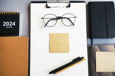 Gri arkaplanda kağıt tutacağı, not defteri, yapışkan notlar, kalemler, gözlükler ve takvimi olan çalışma yeri