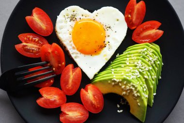 Dilimlenmiş avokado ve vişneli domateslerin yanında kalp şeklinde sahanda yumurta, gri arka planda siyah bir tabakta.