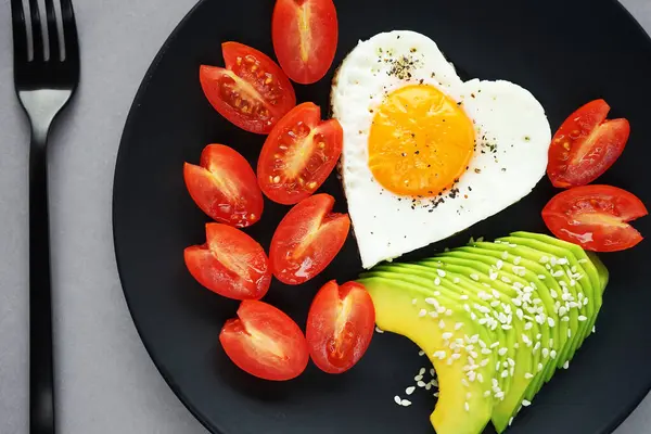 Dilimlenmiş avokado ve vişneli domateslerin yanında kalp şeklinde sahanda yumurta, gri arka planda siyah bir tabakta.