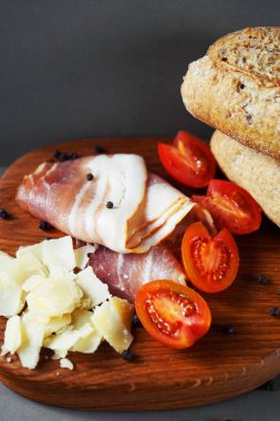 Pastırma dilimleri, parmesan, vişne domatesleri, ekmek ve bıçağın yanında siyah biberler ve gri arka planda.