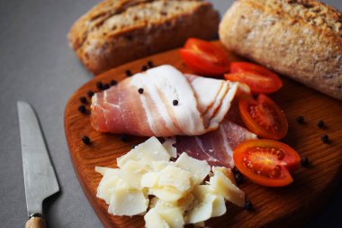 Pastırma dilimleri, parmesan, vişne domatesleri, ekmek ve bıçağın yanında siyah biberler ve gri arka planda.