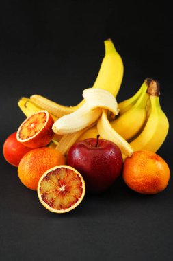 Kırmızı elmanın yanında birkaç olgun sarı muz, koyu arkaplanda portakallar.