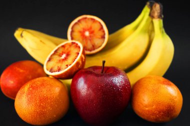 Kırmızı elmanın yanında birkaç olgun sarı muz, koyu arkaplanda portakallar.