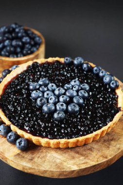 Koyu arkaplanda taze yaban mersininin yanındaki ahşap tezgahta açık yaban mersinli tart.