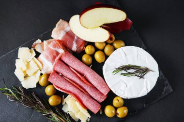 Krem peynirli salam, pastırma, parmesan, zeytin, koyu arkaplanda kırmızı elma ve biberiye.