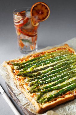 Gri arka planda buzlu limonata bardağının yanında kuşkonmaz ve peynirli turta.