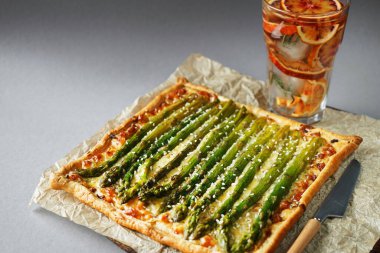 Gri arka planda buzlu limonata bardağının yanında kuşkonmaz ve peynirli turta.