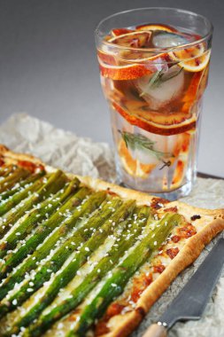 Gri arka planda buzlu limonata bardağının yanında kuşkonmaz ve peynirli turta.