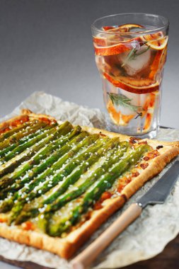 Gri arka planda buzlu limonata bardağının yanında kuşkonmaz ve peynirli turta.